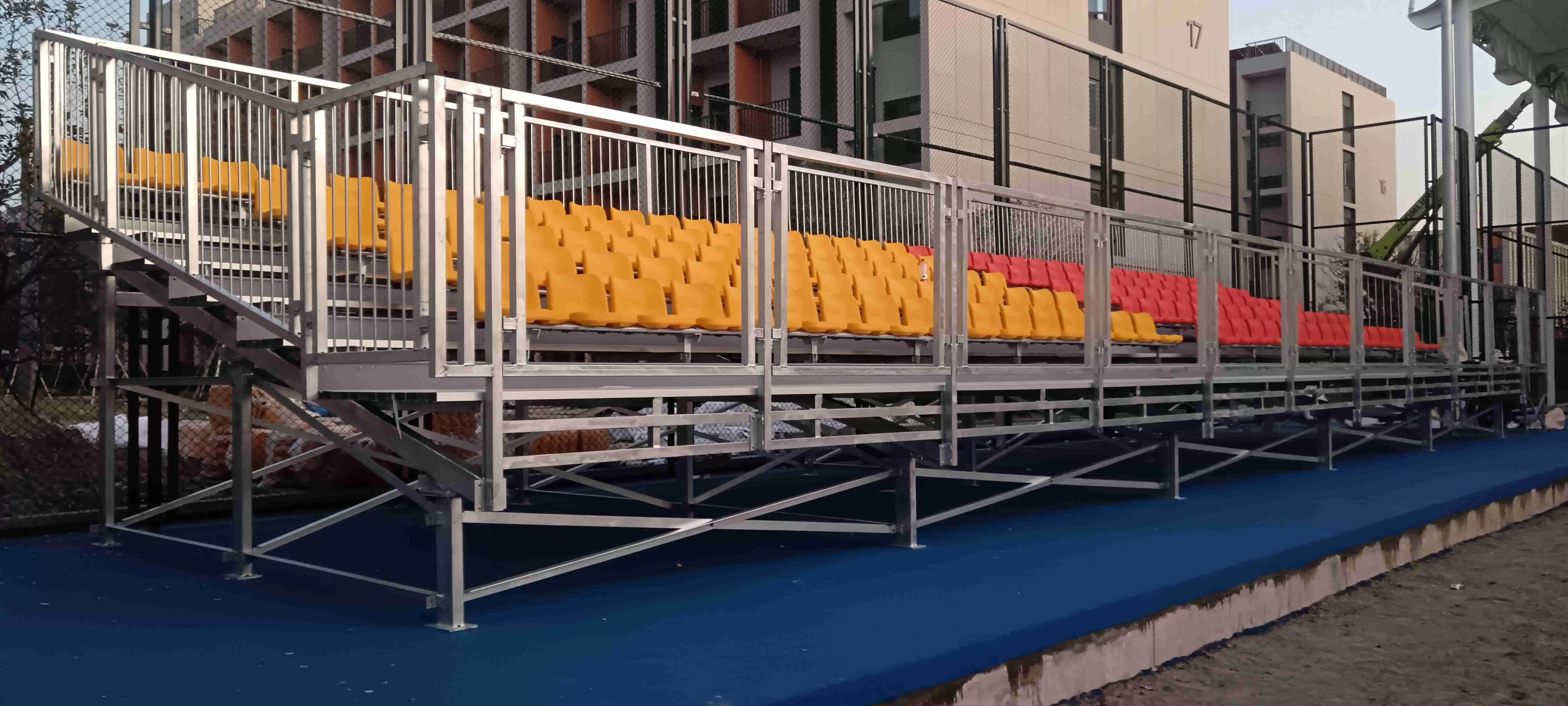 Modular metal bleachers (outdoor metal grandstands) with vibrant yellow/red spectator seating, galvanized metal framework, safety railings, and access staircase—temporary metal grandstands for outdoor sports & event venues