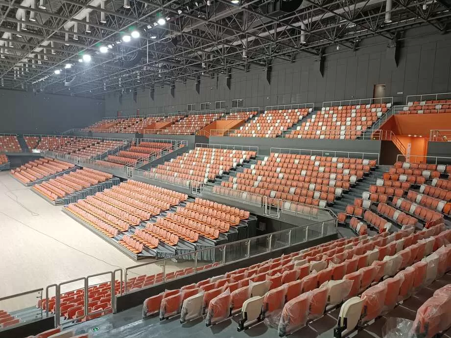 Indoor sports stadium with orange and white telescopic seating, bright lighting, and clear sightlines